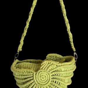 Puede incluir: Un bolso de ganchillo verde lima con un diseño en espiral en la parte delantera. El bolso tiene forma redondeada y una correa larga y trenzada. El bolso está suspendido sobre un fondo negro.