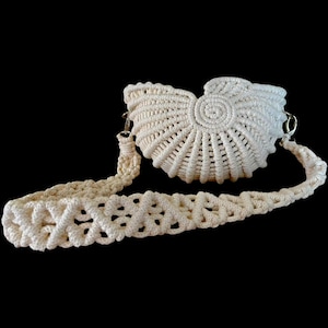 Puede incluir: Bolso de mano de macramé color crema con forma de concha, con una correa larga a juego. El bolso presenta un diseño en espiral en el centro y un patrón de tejido abierto texturizado. La correa también es de macramé, con un diseño calado decorativo y herrajes metálicos.