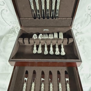 May include: A dark wood flatware chest with a brown velvet interior containing silver-plated utensils. The set includes knives with ornate handles in the lid and drawer, and forks and spoons arranged in the main compartment with tarnish-resistant fabric.