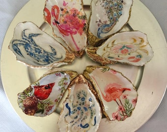 Op de afbeelding: Een verzameling van zeven decoratieve oesterschelpen gerangschikt in een cirkel, elk met unieke decoupage-ontwerpen waaronder flamingo's, een pauw, krabben, een kardinaalvogel en een octopus, allemaal met goudkleurige randen op een ronde schaal.