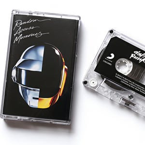 May include: A clear cassette tape and case featuring the album "Random Access Memories" by Daft Punk. The case has a black background with a silver and gold helmet design. The cassette tape is black with white text.