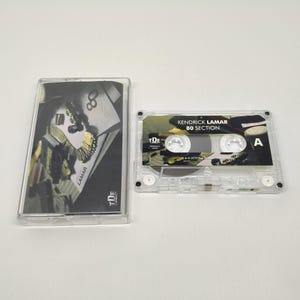 May include: A clear cassette tape with the album title "80 Section" by Kendrick Lamar. The cassette case features a black and white image with gold accents. The "A" side of the tape is visible.