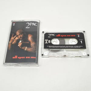 May include: A 2Pac All Eyez On Me cassette tape and its clear plastic case. The cover features a photograph of 2Pac with the text 2PAC and all eyez on me. The cassette itself is clear with a black label featuring the text 2PAC, all eyez on me, and 2.