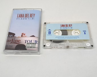 Puede incluir: Una caja de casete de plástico transparente y un casete a juego para el álbum Honeymoon de Lana Del Rey. La carátula muestra un cielo azul claro, una persona con sombrero rosa y un vehículo blanco con el texto Starline Tours.