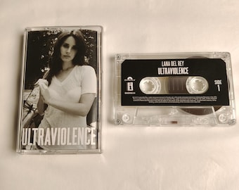 Puede incluir: Una cinta de casete transparente y su estuche con el álbum "Ultraviolence" de Lana Del Rey. El estuche de la cinta muestra una foto en blanco y negro de la artista. La cinta tiene etiquetas negras con texto blanco.