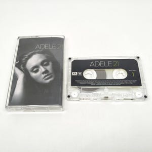 Puede incluir: Una caja de casete de plástico transparente con un retrato en blanco y negro de Adele en la portada con el texto ADELE 21. Al lado hay una cinta de casete transparente con una etiqueta negra, texto blanco que dice ADELE 21 y el número 1.