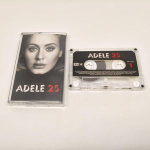 May include: A clear plastic cassette case featuring a black and white portrait of Adele and the text ADELE 25, alongside a matching transparent cassette tape with a black label displaying the text ADELE 25 and the number 1 in red.