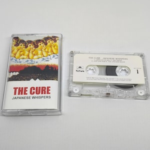 Puede incluir: Una cinta de cassette transparente y su estuche para el álbum "The Cure: Japanese Whispers". El estuche presenta un gráfico de querubines, un paisaje rojo y negro y el nombre de la banda. La cinta de cassette es visible con una etiqueta blanca y la lista de canciones.