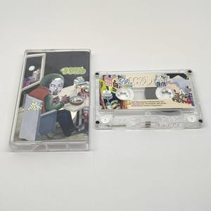 May include: A clear cassette tape and case featuring album art. The case shows a seated figure in a green hood, with the word "DOOM" in green. The cassette itself has the word "DOOM" and colorful city illustrations.