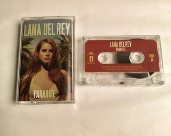 May include: A clear cassette tape and case featuring the text "LANA DEL REY PARADISE". The cassette case has a photo of a woman with long brown hair. The cassette tape is red with white text.