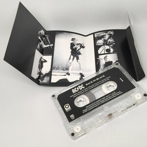 May include: A clear cassette tape with the album title "Back in Black" by AC/DC. The cassette case is transparent, and the label is black and white. The album art is a black and white concert photo. The cassette is on a white surface.