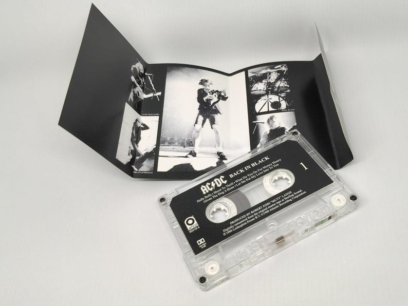 May include: A clear cassette tape with the album title "Back in Black" by AC/DC. The cassette case is transparent, and the label is black and white. The album art is a black and white concert photo. The cassette is on a white surface.