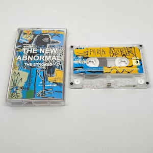 May include: A white ceramic mug with a black handle and the text "But First, Coffee" in a bold, black font. The mug is set against a blurred background, suggesting a cosy setting.