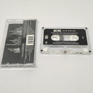 May include: A clear cassette tape case with the cassette tape partially removed. The cassette tape is black with white text that reads "AC/DC BACK IN BLACK". The case is open, revealing the cassette tape's track list.