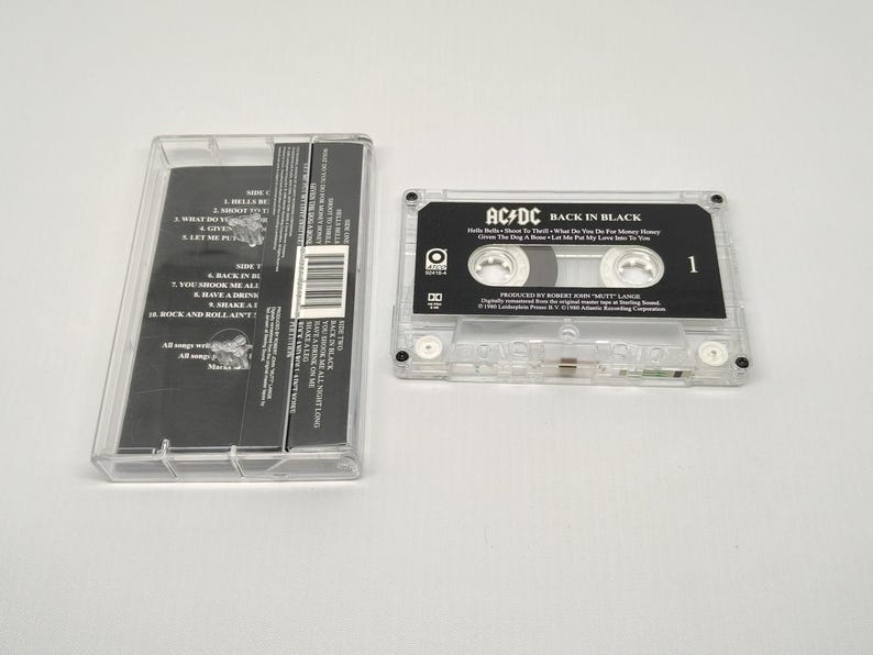 May include: A clear cassette tape case with the cassette tape partially removed. The cassette tape is black with white text that reads "AC/DC BACK IN BLACK". The case is open, revealing the cassette tape's track list.