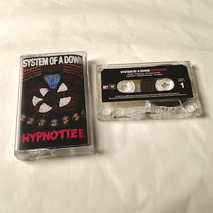 May include: Two clear cassette tapes. One cassette case features the text "SYSTEM OF A DOWN" and "HYPNOTIZE" in red and white. The cassette tape itself has the text "SYSTEM OF A DOWN HYPNOTIZE" and the number "1".