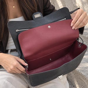 May include: A black leather handbag with a textured exterior and a smooth deep red interior lining. The bag features a rectangular flap with a silver-toned metal clasp closure, a zippered interior pocket, and a snap button fastener.