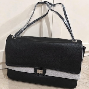 May include: A black textured shoulder bag with a rectangular flap, featuring a silver-toned rectangular twist-lock closure and a light gray felt trim along the bottom edge of the flap. The bag includes a shoulder strap wrapped in clear plastic.