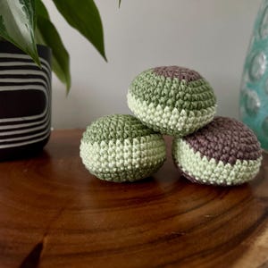 May include: Three crocheted juggling balls in shades of green and brown rest on a wooden surface. The balls are handmade with a textured stitch, and the color scheme includes light green, dark green, and brown.