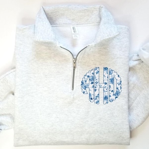 May include: A gray quarter-zip sweatshirt with a blue and white floral monogram on the front. The monogram is in a circle and the letters are "AHD".