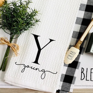 May include: A white kitchen towel with a black "Y" initial and the name "young" in script. A black and white checkered towel is next to it. A wooden spatula with "GRAB" printed on it and a metal pig sign are also visible.