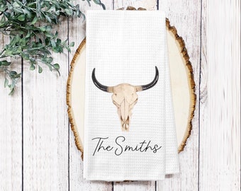 May include: White waffle-weave kitchen towel featuring a watercolor illustration of a bull skull with black horns. The text "The Smiths" is printed in a script font below the skull. The towel is displayed on a wooden slice.