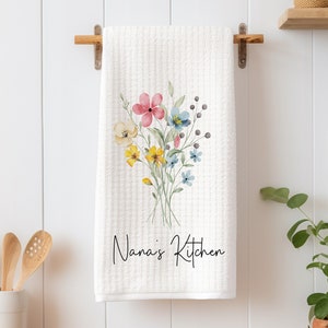 May include: White kitchen towel with a watercolor floral design in shades of pink, yellow, and blue. The text "Nana's Kitchen" is printed in black cursive lettering.
