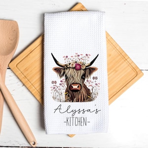May include: White kitchen towel with a floral print of a Highland cow with a flower crown. The text "Alyssa's Kitchen" is printed below the cow.