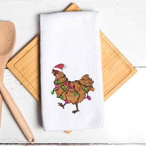 May include: A white kitchen towel with a brown chicken wearing a red Santa hat and wrapped in colorful Christmas lights.