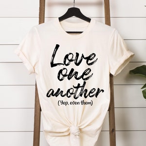 May include: A white t-shirt with black text that reads "Love one another (Yep, even them)".