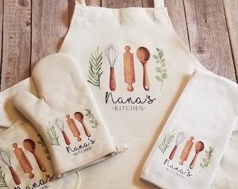 Puede incluir: Un juego de textiles de cocina con fondo blanco y un diseño de acuarela marrón y verde de utensilios de cocina y vegetación. El juego incluye un delantal, un agarrador de ollas, un guante para horno y una toalla de té. El texto "Nana's Kitchen" está impreso en cada artículo.