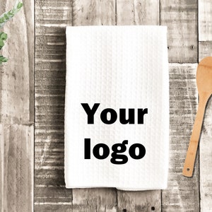 May include: White kitchen towel with the text "Your logo" printed in black on a wooden background with a wooden spoon.