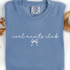 May include: A folded, light blue t-shirt with the words "cool aunts club" in white script, along with a bow graphic. The shirt has a crew neck and short sleeves. A small tag is visible at the neckline.