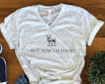 May include: A heather gray V-neck t-shirt with a graphic of a lamb and the text "BUT NOW I'M FOUND". The shirt is paired with blue denim shorts, navy blue sneakers, and accessories.