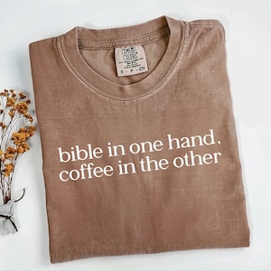 Pu&ograve; includere: Una maglietta Comfort Colors marrone con la scritta bianca "bible in one hand, coffee in the other". La maglietta ha un girocollo e maniche corte. Un piccolo rametto di fiori secchi &egrave; a sinistra della maglietta.