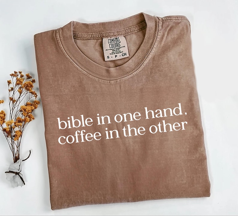 Pu&ograve; includere: Una maglietta Comfort Colors marrone con la scritta bianca "bible in one hand, coffee in the other". La maglietta ha un girocollo e maniche corte. Un piccolo rametto di fiori secchi &egrave; a sinistra della maglietta.
