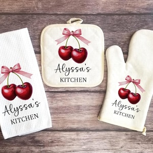 May include: A set of three kitchen towels with a cherry and bow design. The towels are white with pink bows and red cherries. The text "Alyssa's Kitchen" is printed on each towel.