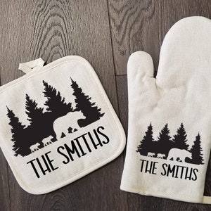 May include: A set of two white cotton oven mitts with black bear and pine tree silhouettes. The mitts are personalized with the text "The Smiths".
