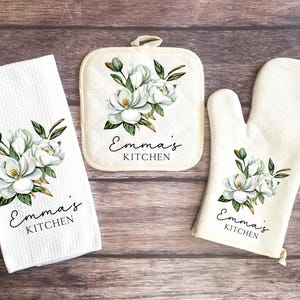 May include: A white kitchen towel, pot holder, and oven mitt set. Each item features a watercolor floral design with white flowers and green leaves. The text "Emma's Kitchen" is printed on each item.