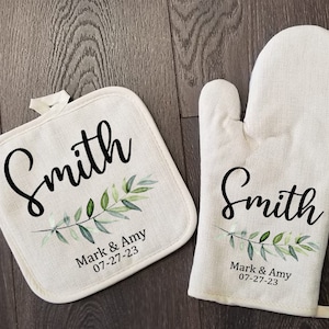 May include: A matching pot holder and oven mitt set. Both are off-white with the name "Smith" in black script, a green and blue leaf design, and the text "Mark & Amy 07-27-23".