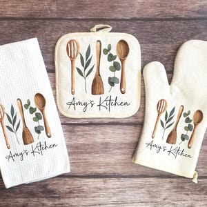 May include: Set of three kitchen towels with a white background and green leaves and wooden kitchen utensils. The towels have the text "Amy's Kitchen" printed on them.