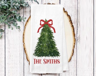 May include: White waffle-weave kitchen towel featuring a green Christmas tree design with a red bow at the top. The words "THE SMITHS" are printed in red at the bottom of the towel. The towel is displayed on a wooden slice.