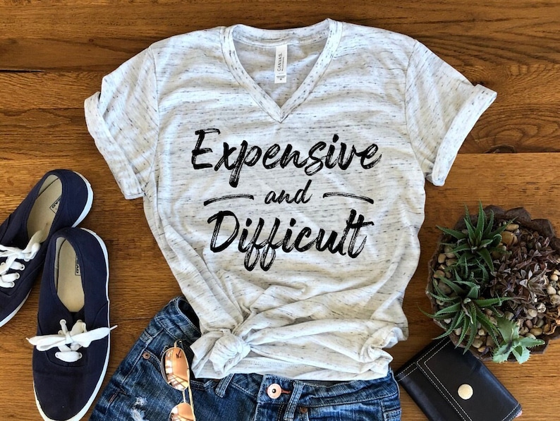 以下が含まれることがあります： 白地に斑点模様のVネックTシャツ。黒い文字で「Expensive and Difficult」と書かれている。
