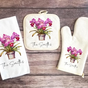 May include: A white kitchen towel, pot holder, and oven mitt set. Each item features a watercolor illustration of a potted orchid with pink and purple flowers. The text "The Smiths" is printed below the floral design.