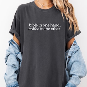Pu&ograve; includere: T-shirt grigio scuro con la scritta bianca "bible in one hand, coffee in the other". La maglietta ha maniche corte e una vestibilit&agrave; rilassata. Sotto &egrave; indossata una giacca di jeans azzurra.