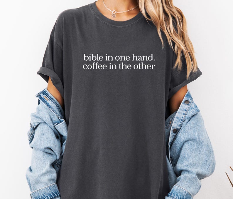 Pu&ograve; includere: T-shirt grigio scuro con la scritta bianca "bible in one hand, coffee in the other". La maglietta ha maniche corte e una vestibilit&agrave; rilassata. Sotto &egrave; indossata una giacca di jeans azzurra.