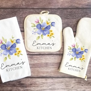 May include: A white kitchen towel, pot holder, and oven mitt set. Each item features a watercolor floral design with blue, yellow, and pink flowers. The text "Emma's KITCHEN" is printed on each item.