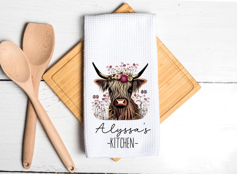 May include: White kitchen towel with a floral print of a Highland cow with a flower crown. The text "Alyssa's Kitchen" is printed below the cow.