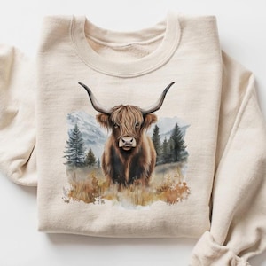 May include: A beige sweatshirt featuring a watercolor illustration of a Highland cow. The cow has long, curved horns and shaggy brown fur. The background includes a mountain range and evergreen trees, with a grassy foreground.