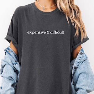 May include: Dark gray t-shirt with the text "expensive & difficult" in white. The shirt has a crew neck and short sleeves. A denim jacket is worn over the shirt.